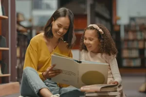 Adult and child sitting on the floor in a library reading a large book together with stacked books nearby